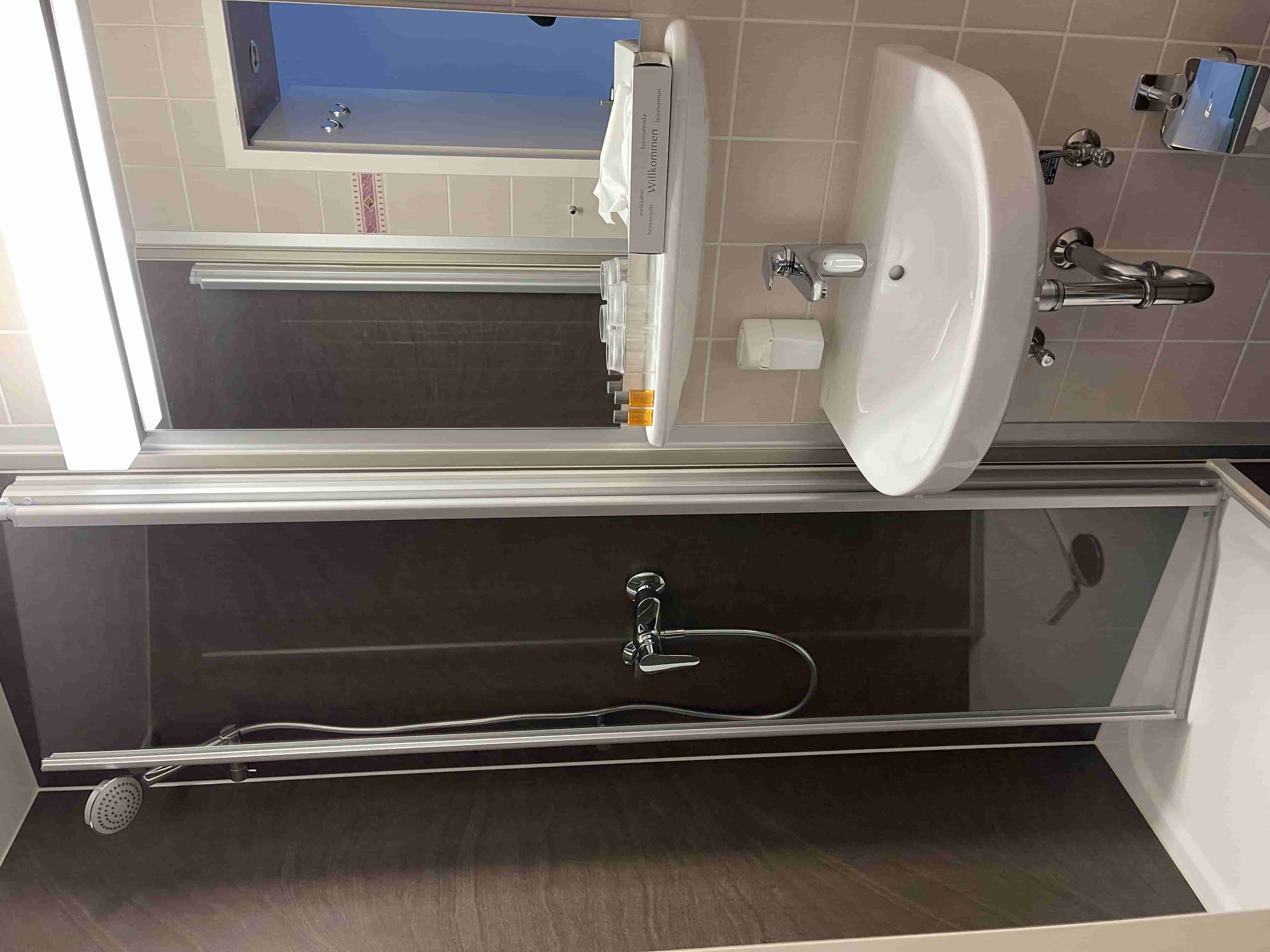 Kleines Badezimmer mit Duschecke, Spiegel, Waschbecken, Seifenspender und Toilettenpapierhalter.