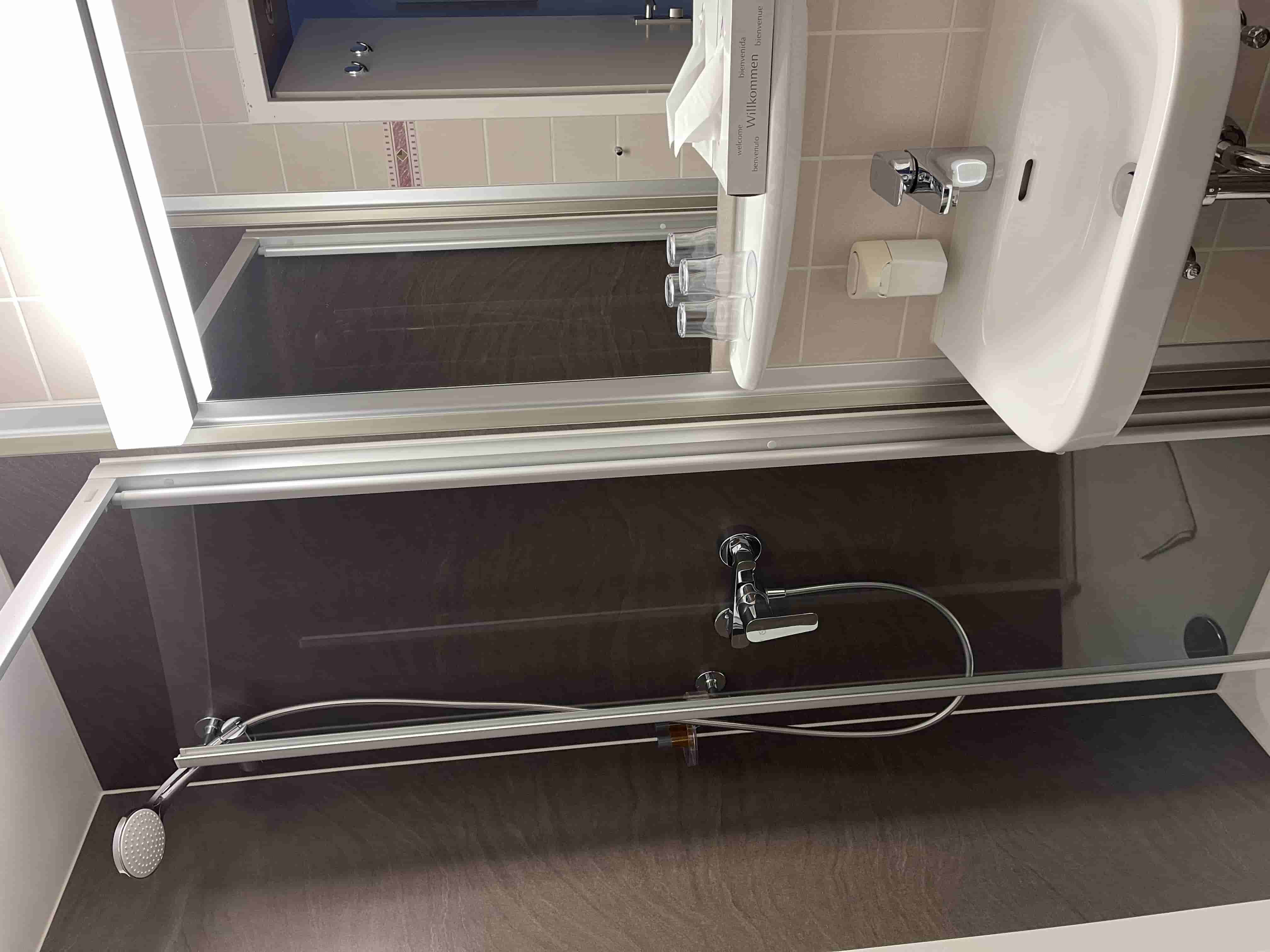 Kleine Hotelbadezimmer-Dusche mit Glasduschwand, weißem Waschbecken, Wandspiegel, Seifenspender und mehreren umgedrehten Trinkgläsern.