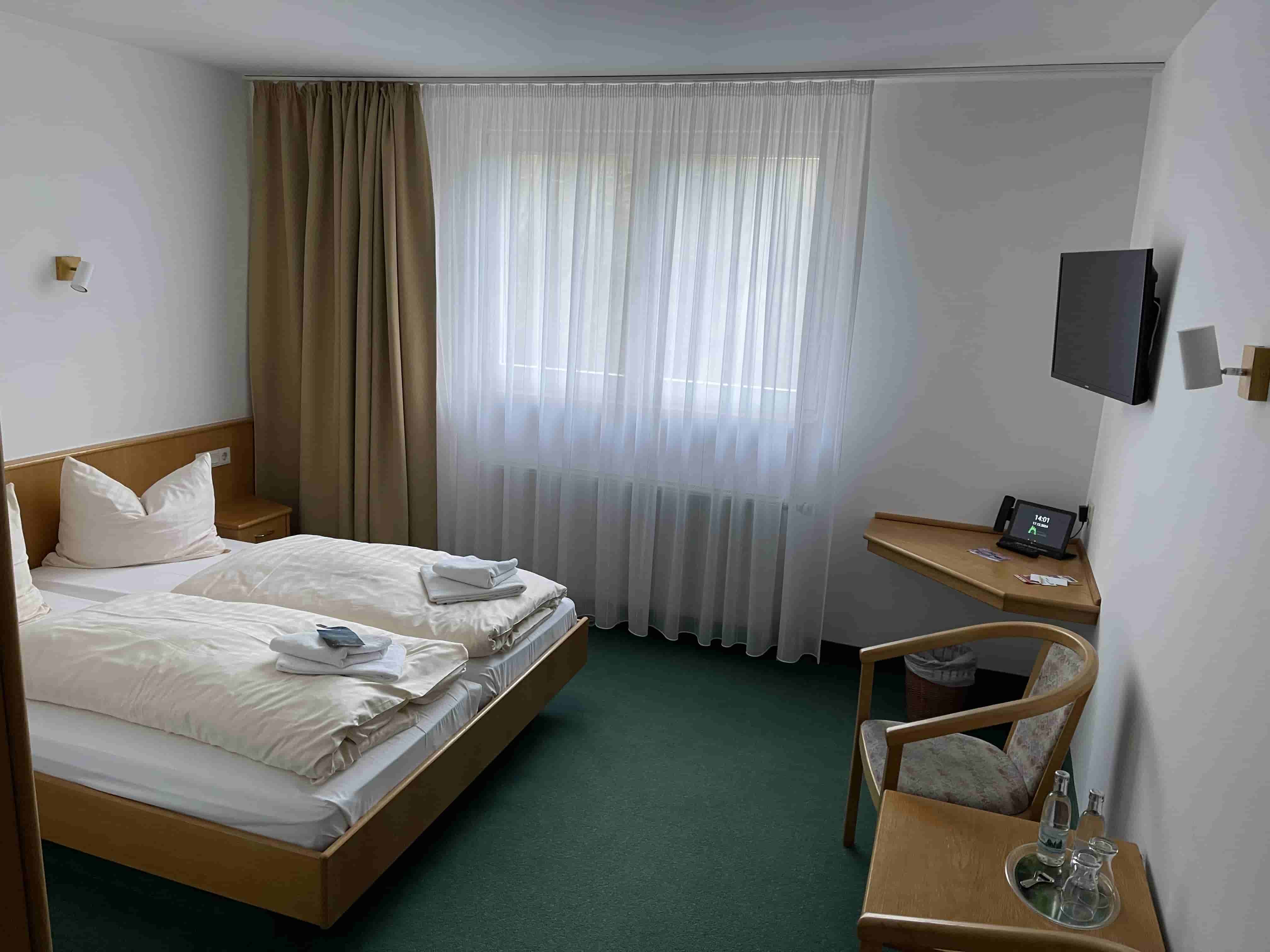 Kompaktes Hotelzimmer mit zwei Einzelbetten, grünem Teppich, Fenster mit Vorhängen, Wandfernseher und kleinem Tisch mit Stuhl.