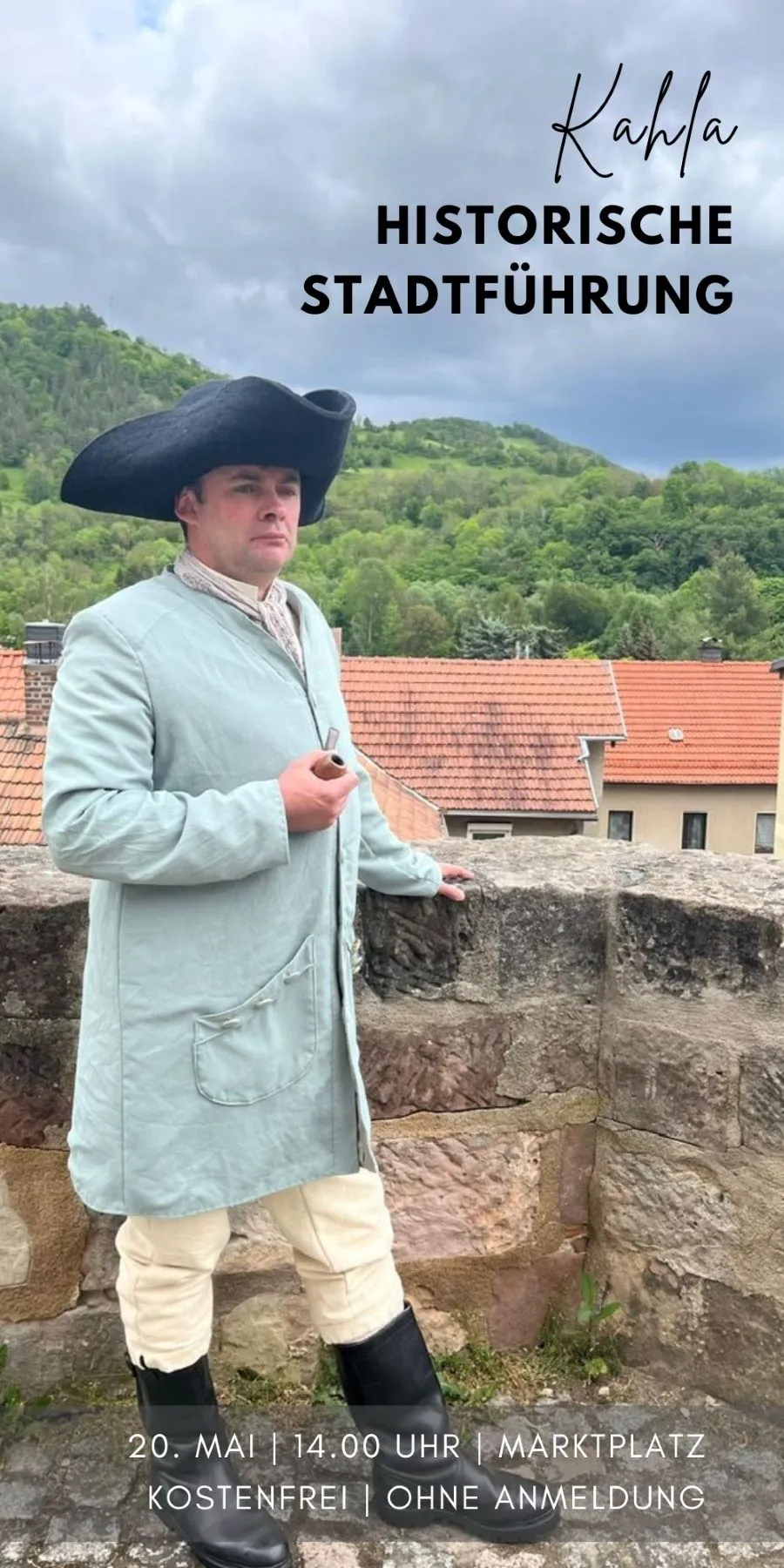 Mann in historischer Kleidung mit Dreispitz und Pfeife steht vor einer Steinmauer mit Häusern und bewaldeten Hügeln im Hintergrund.