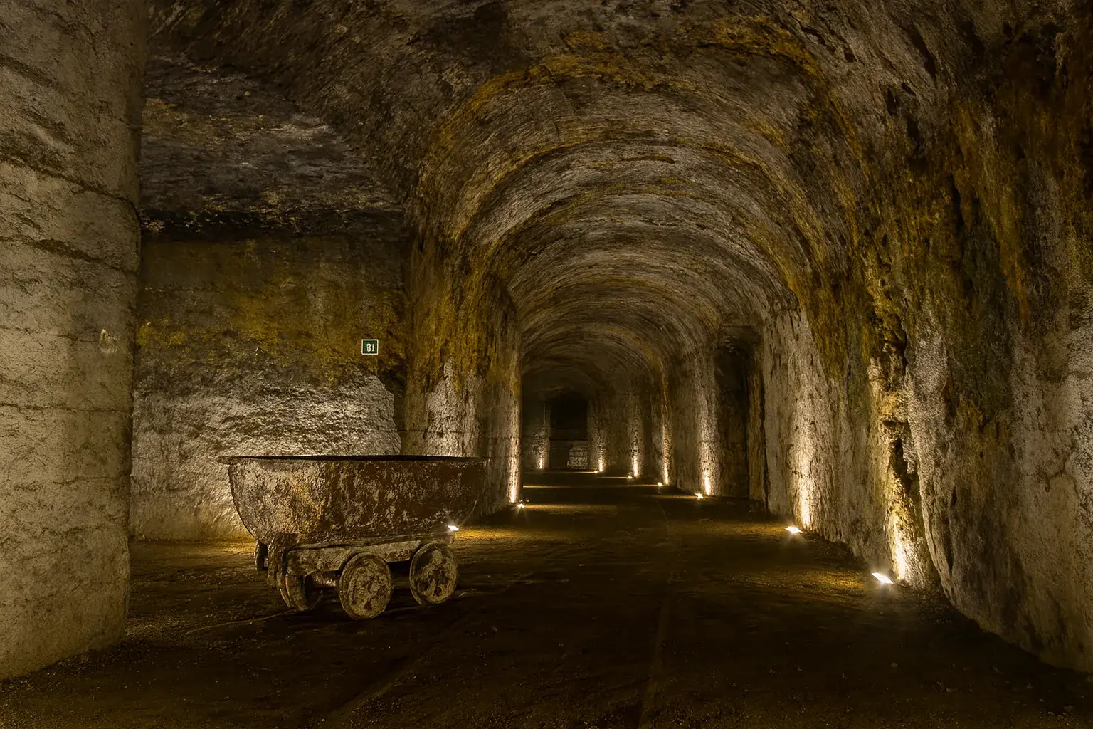 Beleuchteter Kieswagen auf Schienen in einem langen, gewölbten Bergwerkstunnel aus Stein.