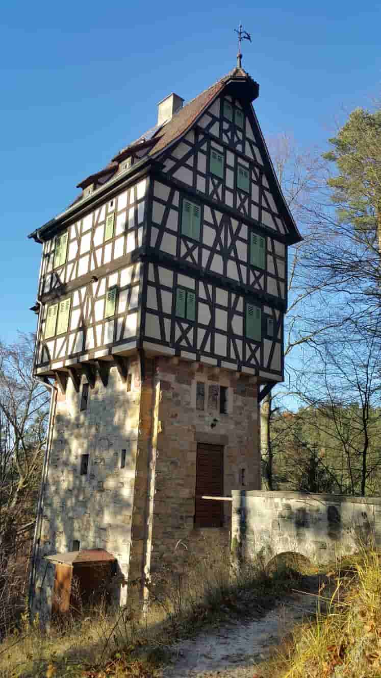 Historischer Fachwerkturm aus Stein und Holz mit grünen Fensterläden, umgeben von Bäumen unter blauem Himmel.