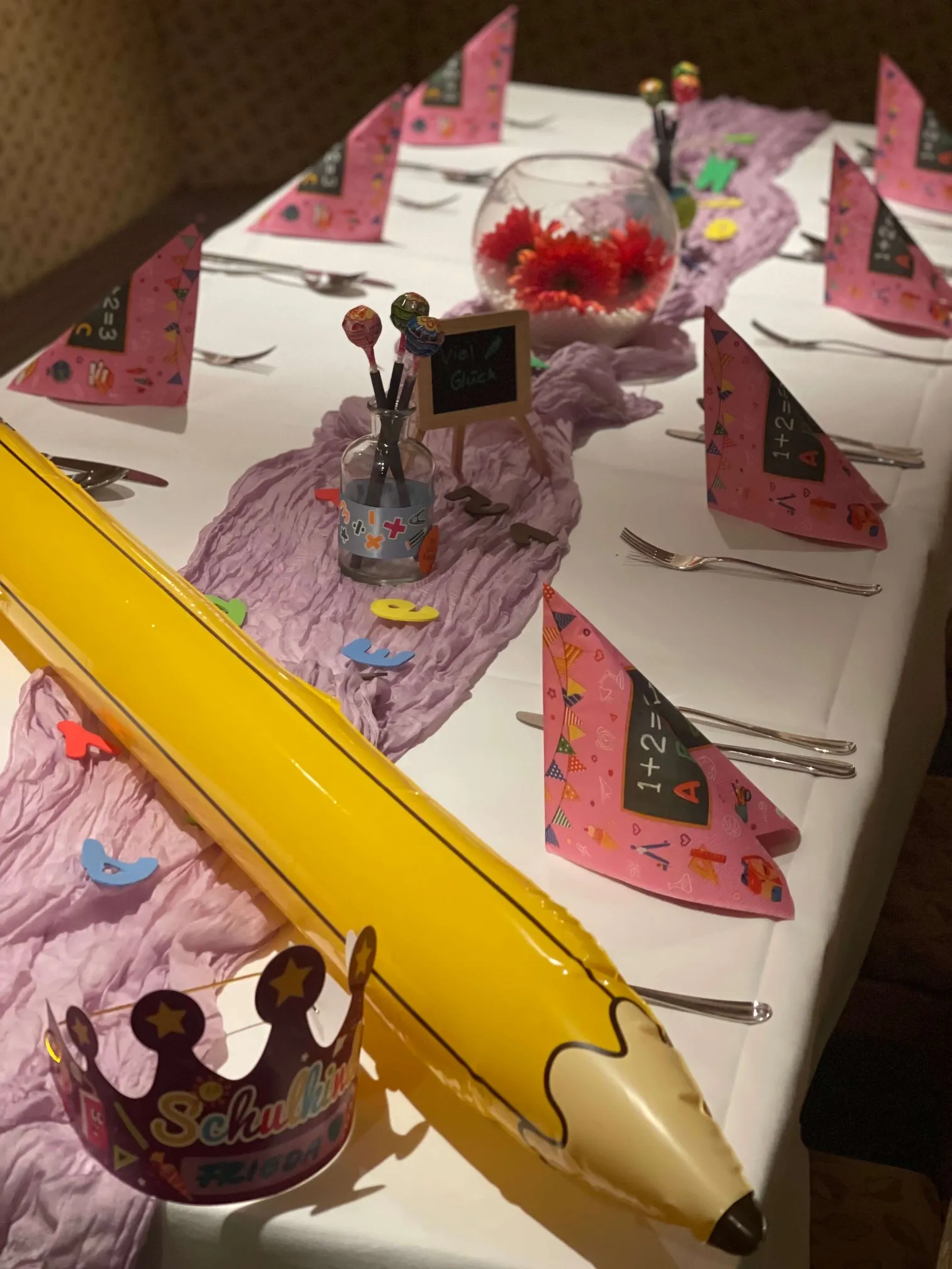 Festlich gedeckter Tisch mit rosa Servietten, einem gelben Riesenbleistift, einer Krone mit der Aufschrift 'Schulkind' und Dekoration für eine Einschulungsfeier.