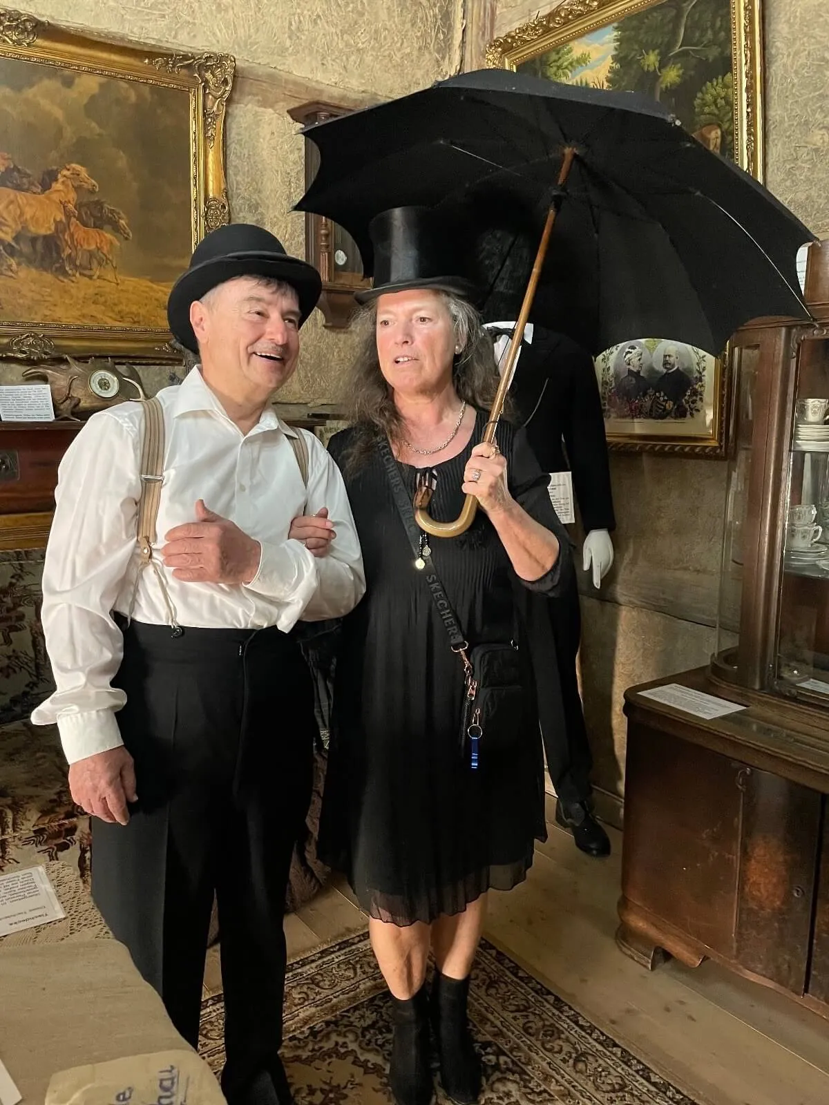 Ein Mann und eine Frau in historischen Kostümen mit Hut; die Frau hält einen schwarzen Regenschirm in einem gemütlich eingerichteten Raum mit Gemälden und Möbeln.