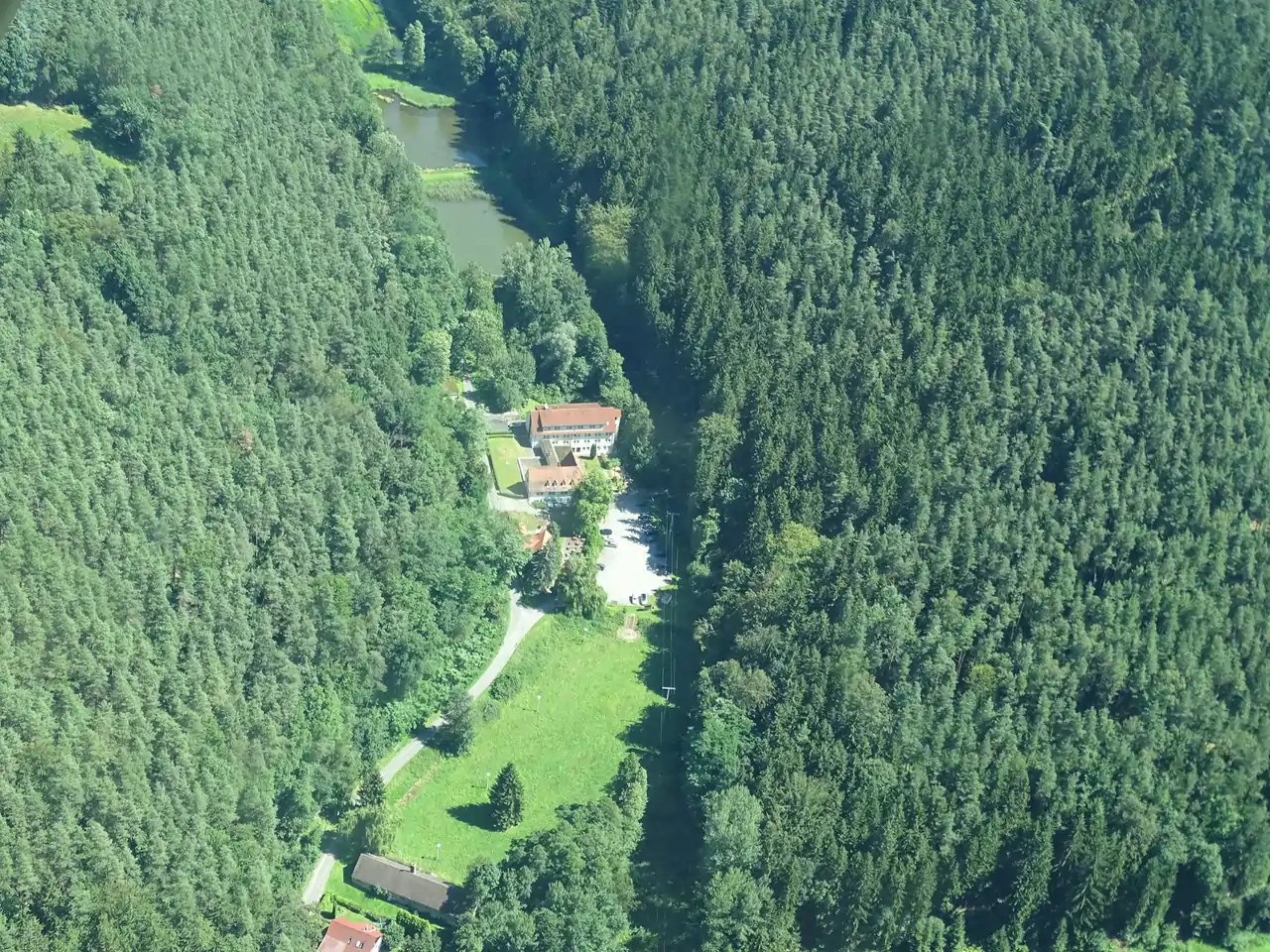 Luftaufnahme eines Hauses mit roten Dächern, umgeben von dichtem grünen Wald und einem kleinen Teich.