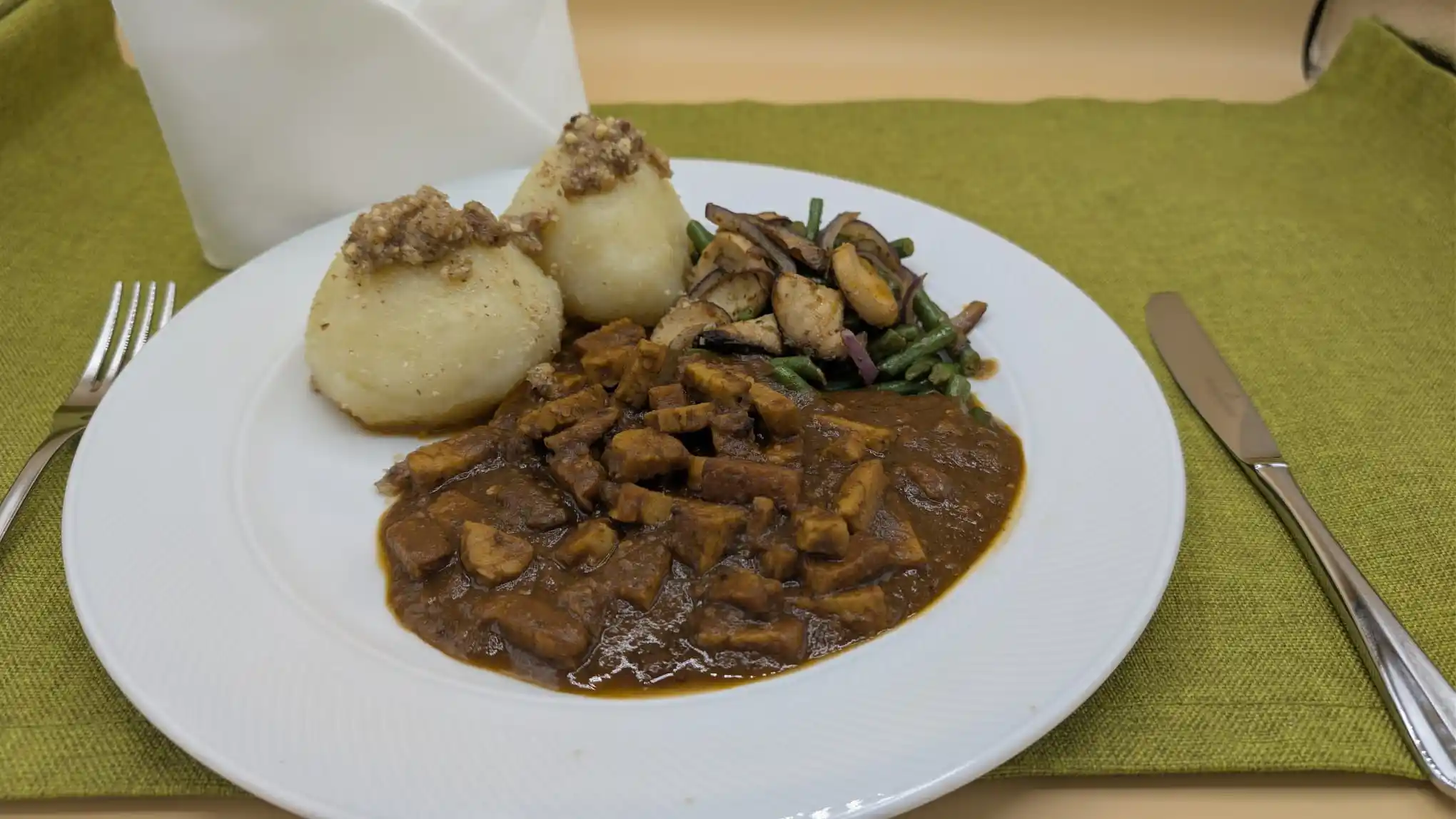 Teller mit Gulasch, Kartoffelknödeln mit Zwiebelbröseln und gebratenem Gemüse auf grünem Tischset.