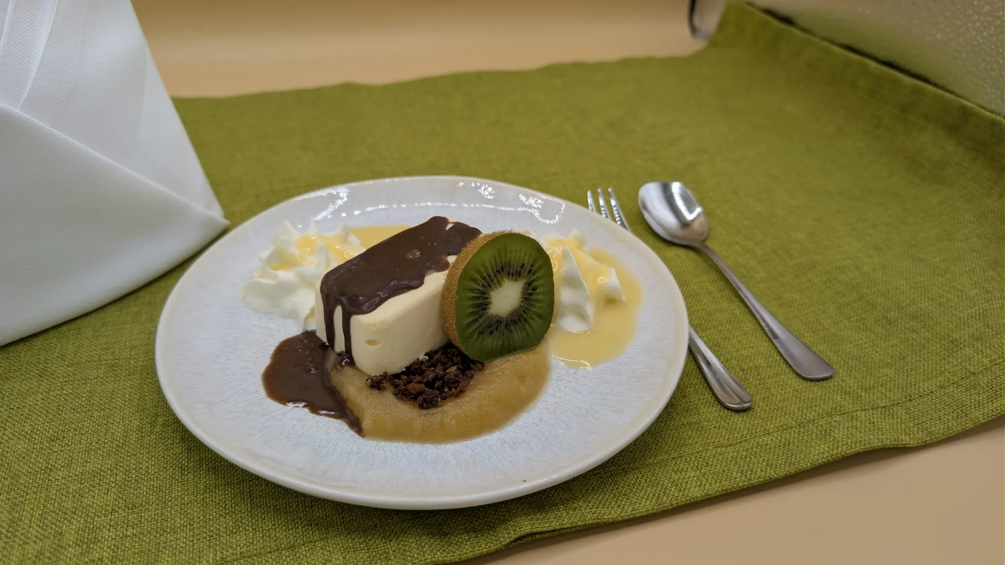 Dessert with cream, kiwi slice, chocolate sauce, and whipped cream on a white plate on green cloth.